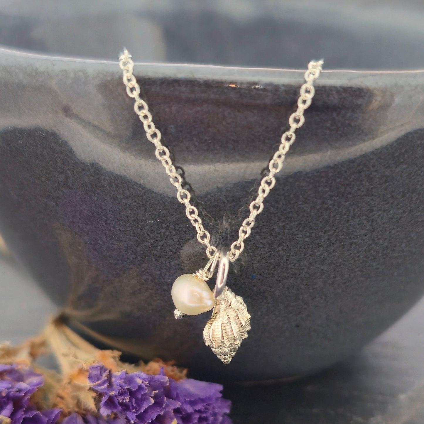A silver periwinkle shell pendant with a cream coloured pearl suspended from a silver chain. On a black bowl with purple flowers. Handmade. Mijoux Jewellery.
