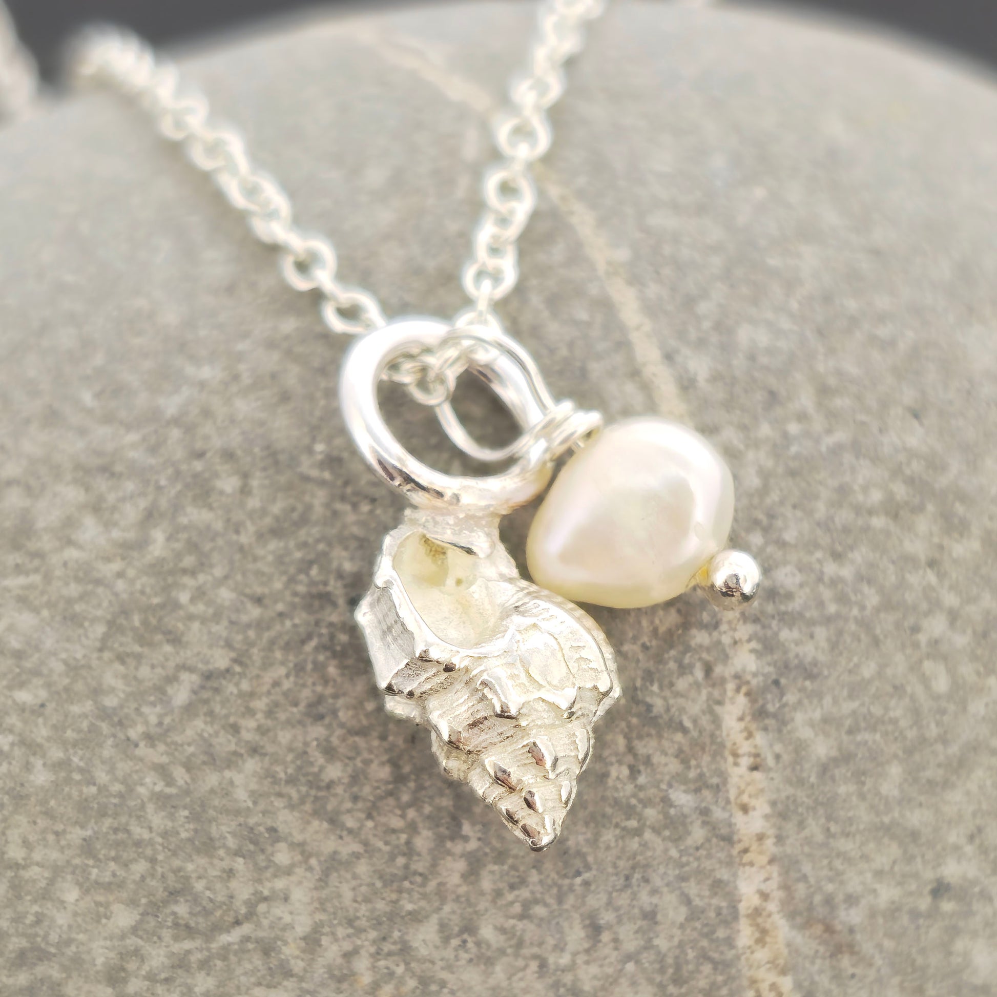 A silver periwinkle shell pendant with a cream coloured pearl suspended from a silver chain. On a stone background. Handmade. Mijoux Jewellery.
