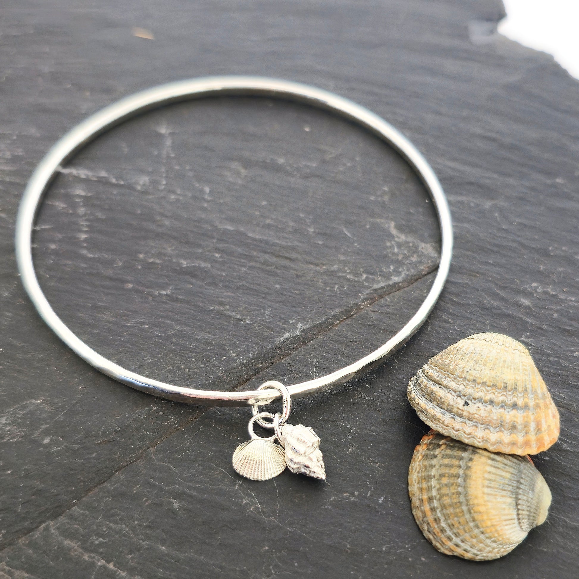 A round silver bangle with 2 seashell charms. 1 cockle shell and 1 periwonkle shell. On a dark background with real shells. Handmade, Mijoux Jewellery.
