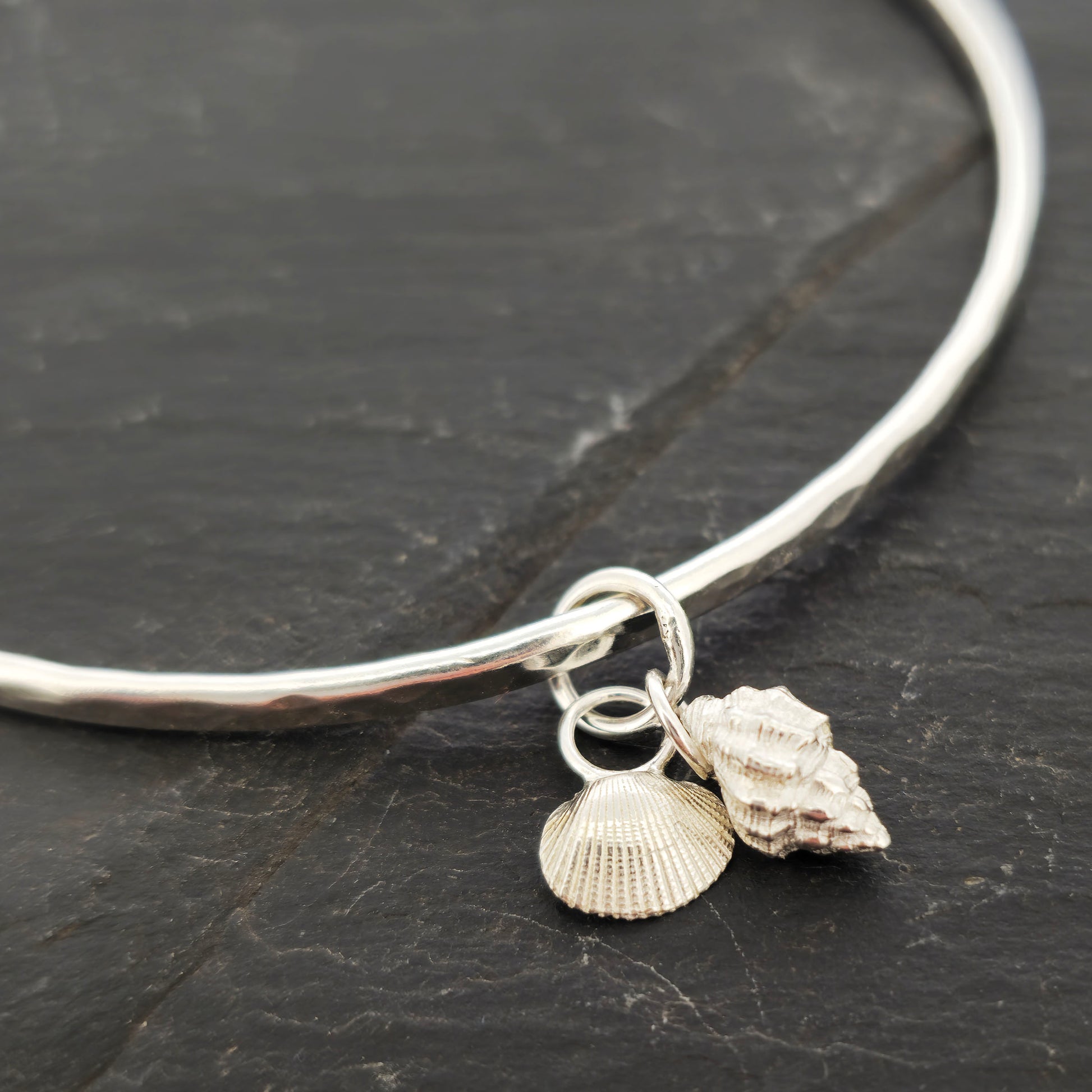 A round silver hammered bangle with 2 seashell charms. 1 cockle shell and 1 periwonkle shell. On a dark background. Handmade, Mijoux Jewellery.