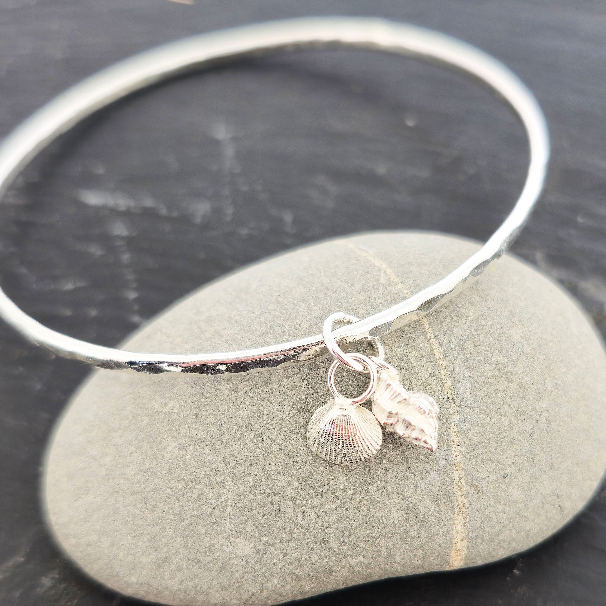 A round hammered silver bangle with 2 seashell charms. 1 cockle shell and 1 periwonkle shell. On a dark background with a pebble. Handmade, Mijoux Jewellery.
