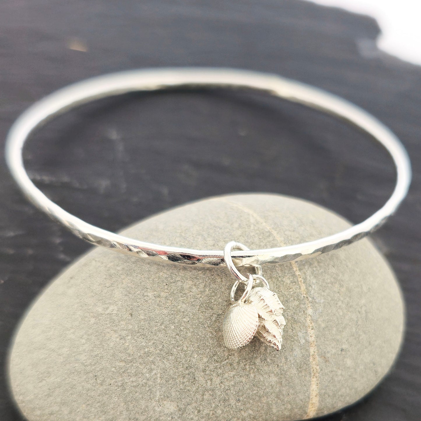 A round silver hammered bangle with 2 seashell charms. 1 cockle shell and 1 periwonkle shell. On a dark background a pebble. Handmade, Mijoux Jewellery.
