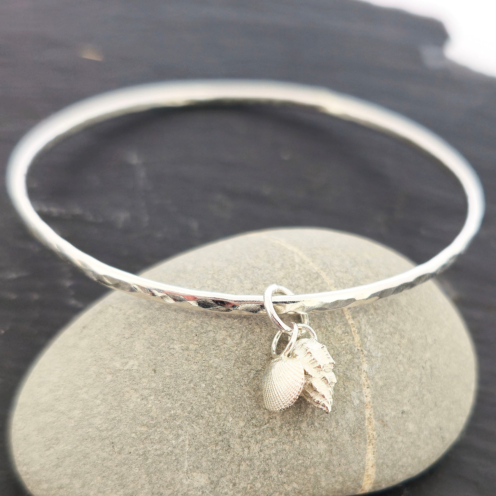 A round silver hammered bangle with 2 seashell charms. 1 cockle shell and 1 periwonkle shell. On a dark background a pebble. Handmade, Mijoux Jewellery.