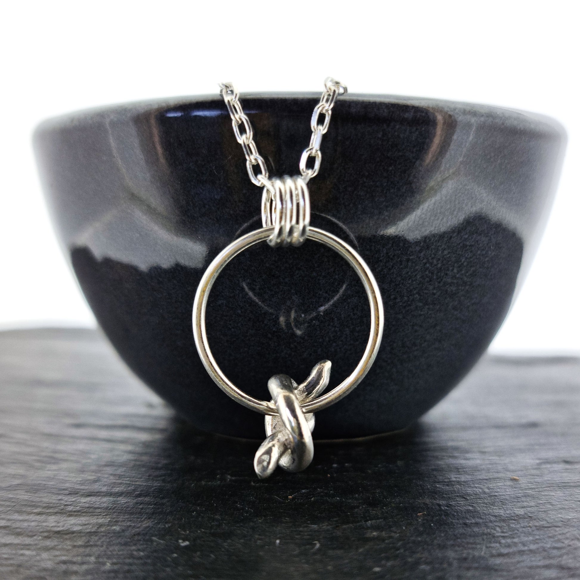 Silver circle pendant with spinning knot charm and triple circle bail. Suspended on a silver chain. Pictured on a black bowl.