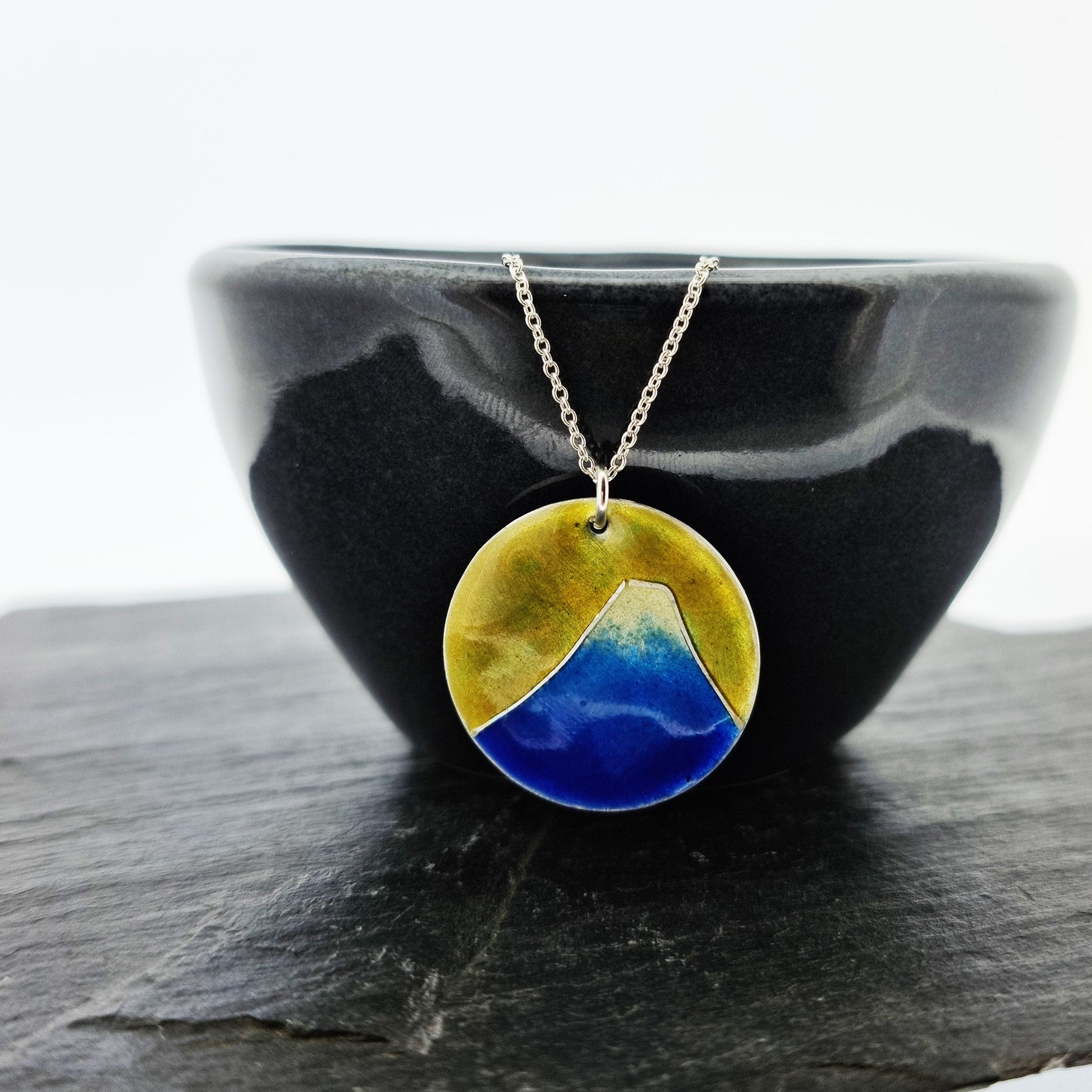 Round silver pendant with a mountain on in graduated blue enamel and a white top and a yellow enamel background. Suspended from a silver chain - on a bowl