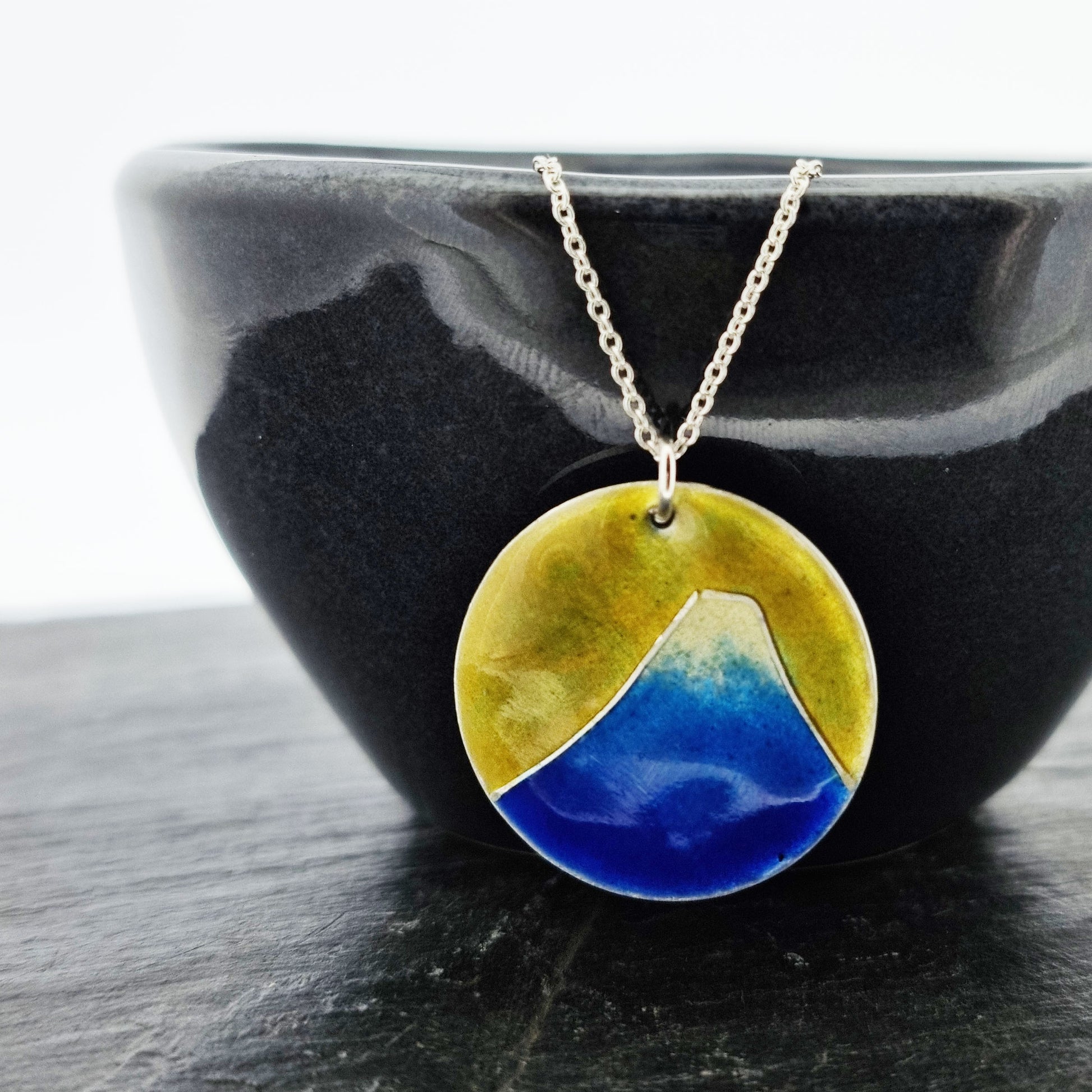 Round silver pendant with a mountain on in graduated blue enamel and a white top and a yellow enamel background. Suspended from a silver chain - on a bowl