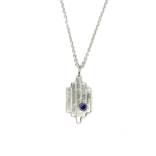 A silver Art Deco style pendant with 5 lines and a dark blue sapphire set off-centre. On a silver chain.