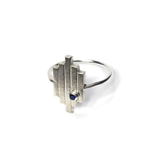 A silver Art Deco style ring with 5 lines and an off-centre blue sapphire gemstone.