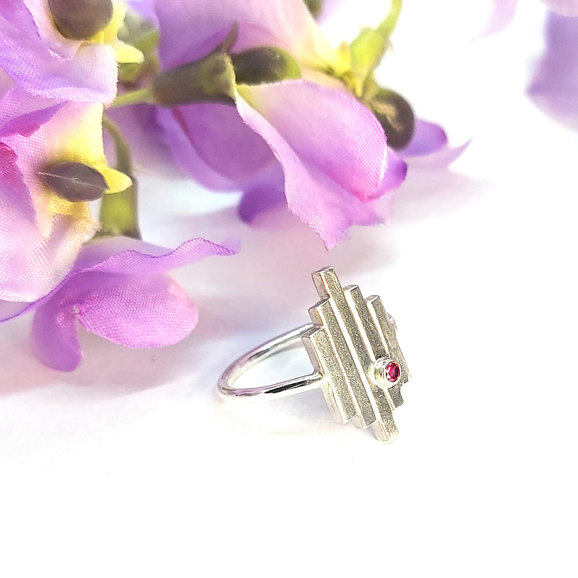 An Art Deco style silver ring with 5 straight lines and an off-centre pinky red ruby. Pictured with flowers.