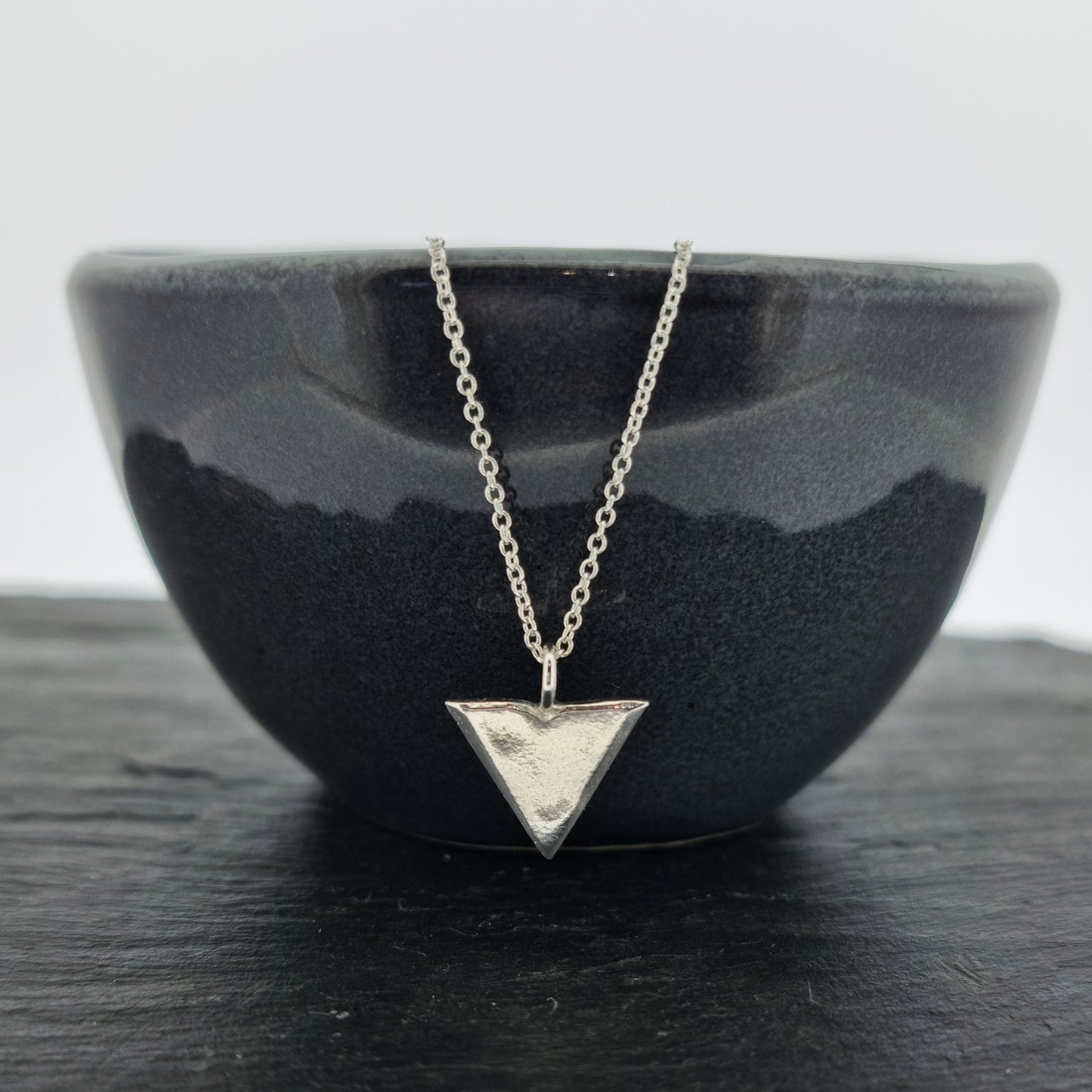 A silver beveled triangle shaped pendant on a silver chain. Hanging from black bowl.