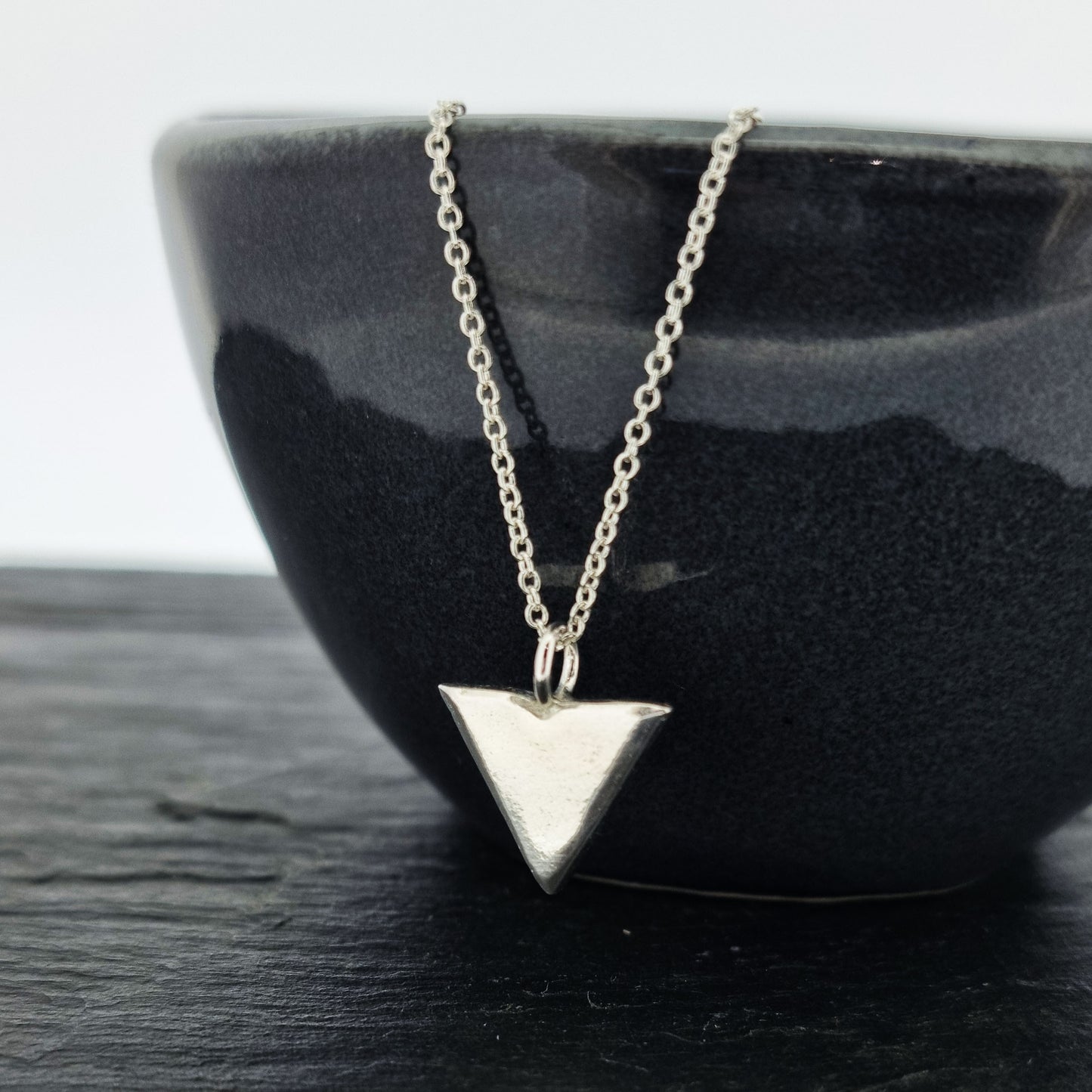 A silver beveled triangle shaped pendant on a silver chain. Hanging from black bowl.