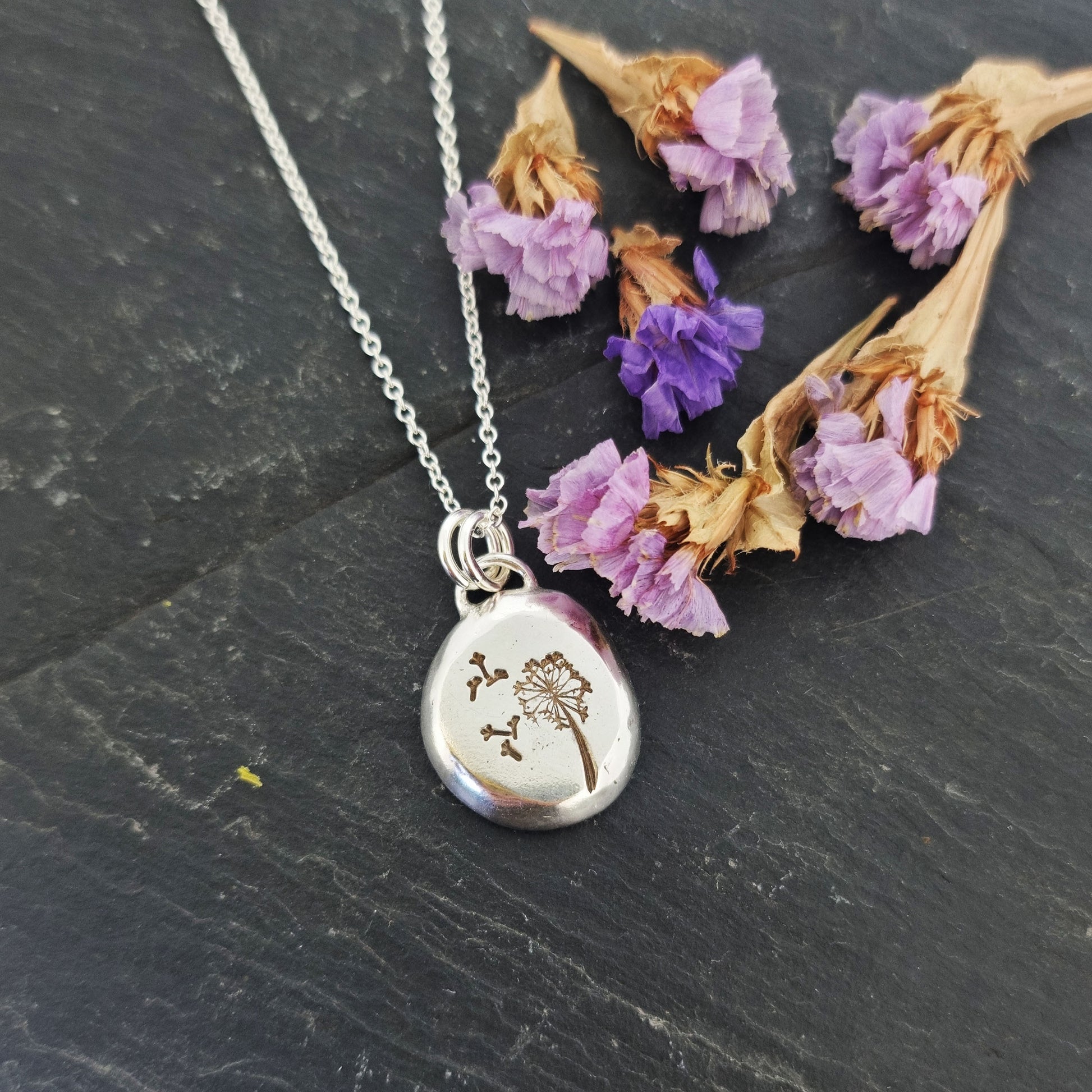 A silver organic round flat pebble pendant with dandelion and fluff engraved on it. On a silver chain. Shown on slate with flowers.