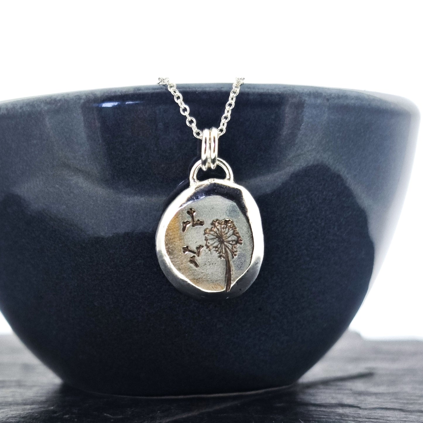 A silver organic round flat pebble pendant with dandelion and fluff engraved on it. On a silver chain. Shown on a black bowl.