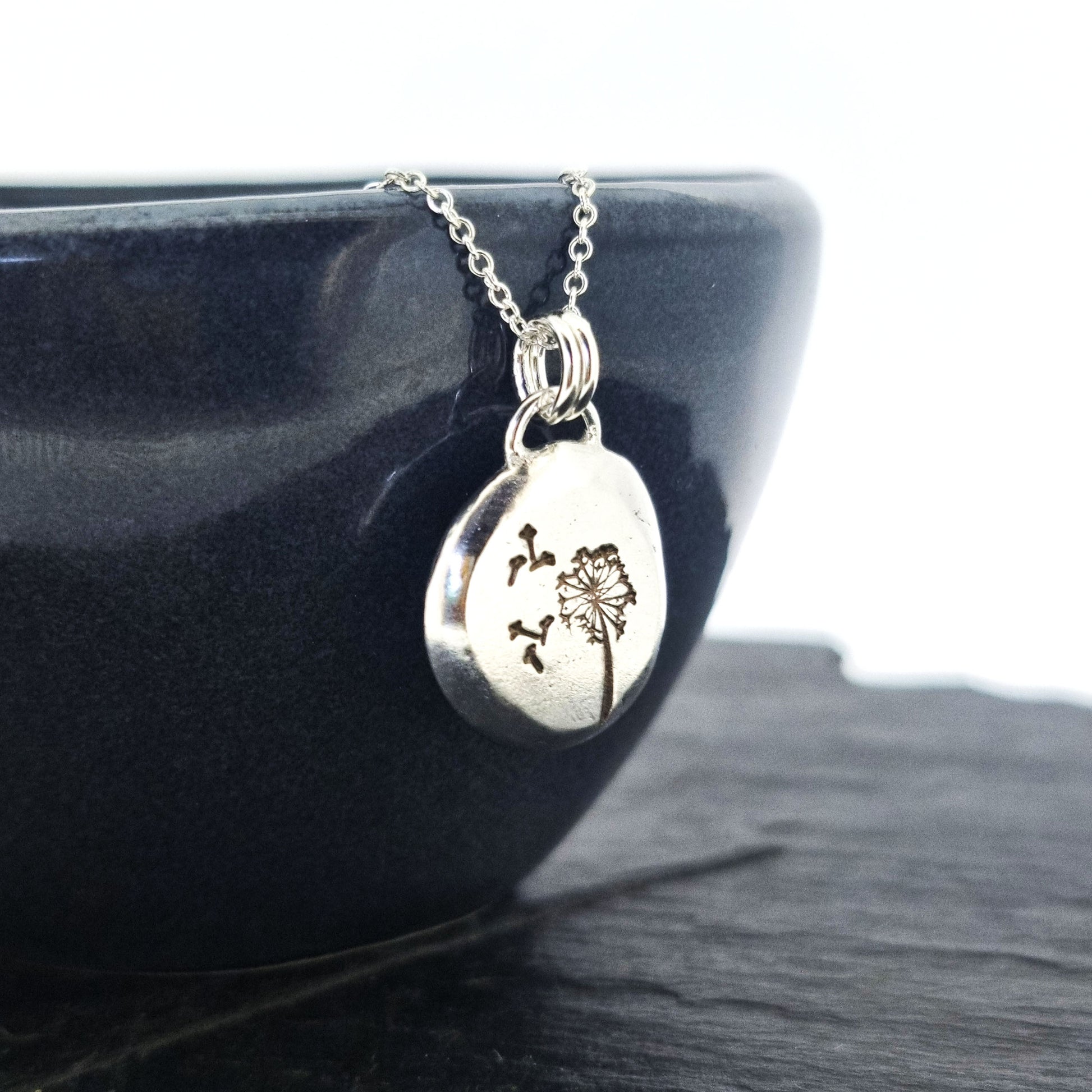 A silver organic round flat pebble pendant with dandelion and fluff engraved on it. On a silver chain. Shown to the side on a black bowl.