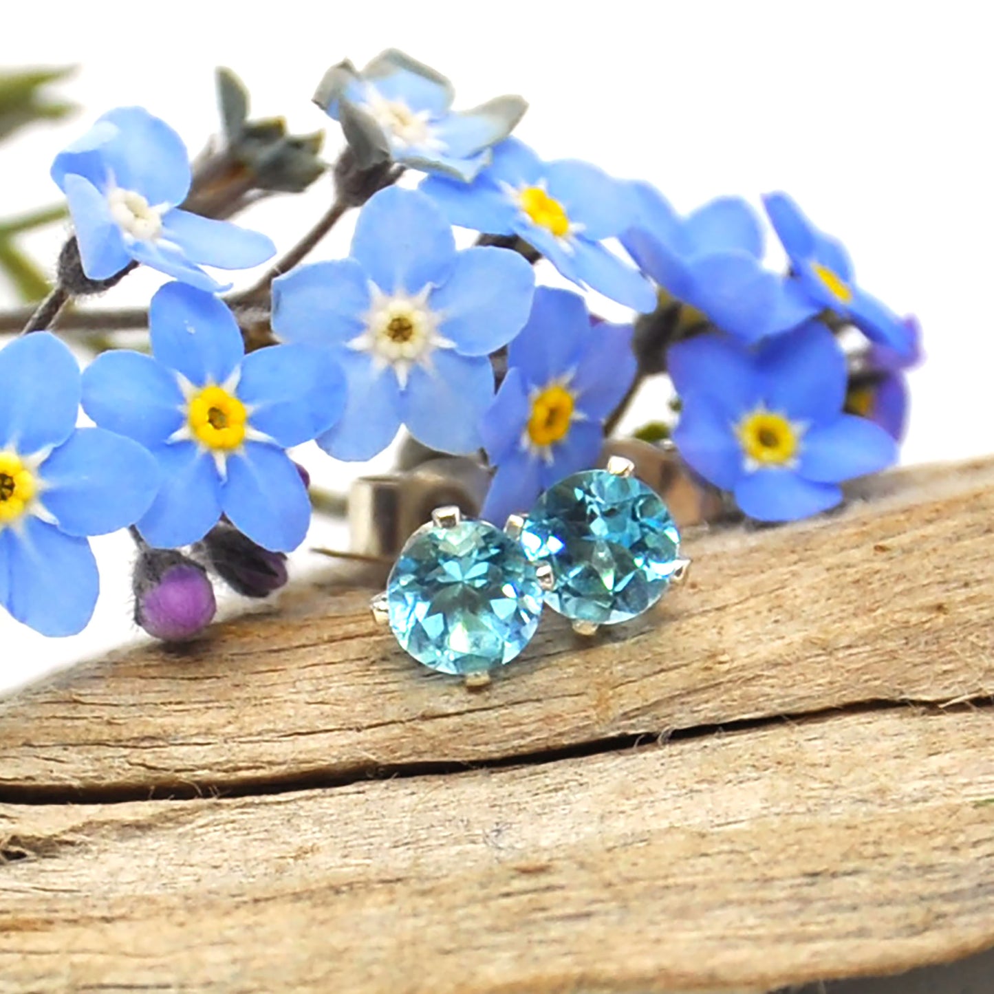 Silver 4 claw stud earrings with bright blue topaz gemstones. Pictured with flowers.