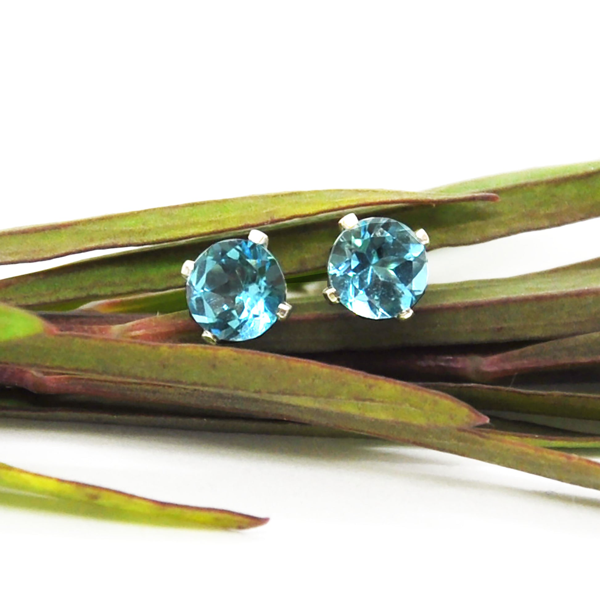Silver 4 claw stud earrings with bright blue topaz gemstones. Pictured with leaves.