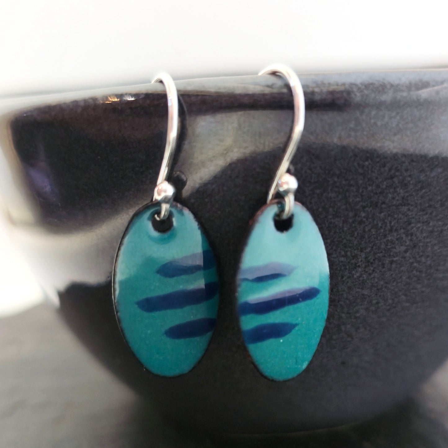 Handmade oval drop earrings with a teal green background and dark blue stripes. On silver ear hooks. Shown on a dark bowl. Mijoux Jewellery.
