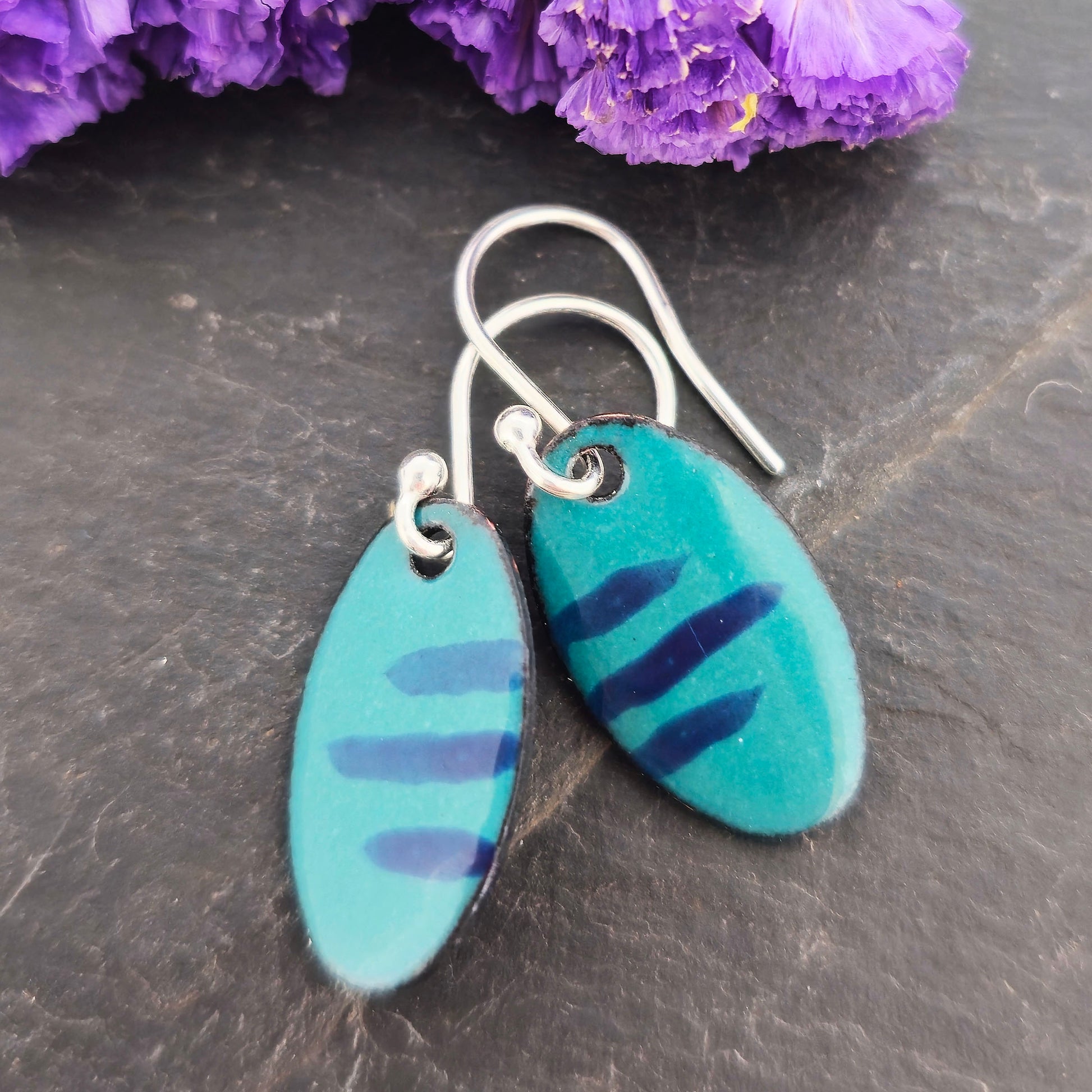 Handmade oval drop earrings with a teal green background and dark blue stripes. On silver ear hooks. Shown on a dark background with purple flowers. Mijoux Jewellery.