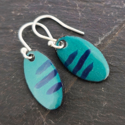 Handmade oval drop earrings with a teal green background and dark blue stripes. On silver ear hooks. Shown on a dark background. Mijoux Jewellery.