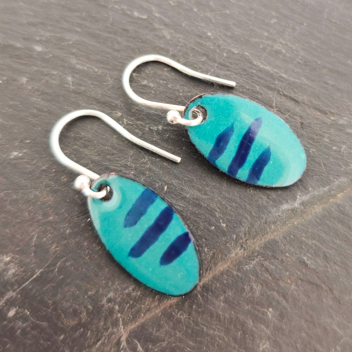 Handmade oval drop earrings with a teal green background and dark blue stripes. On silver ear hooks. Shown on a dark background. Mijoux Jewellery.