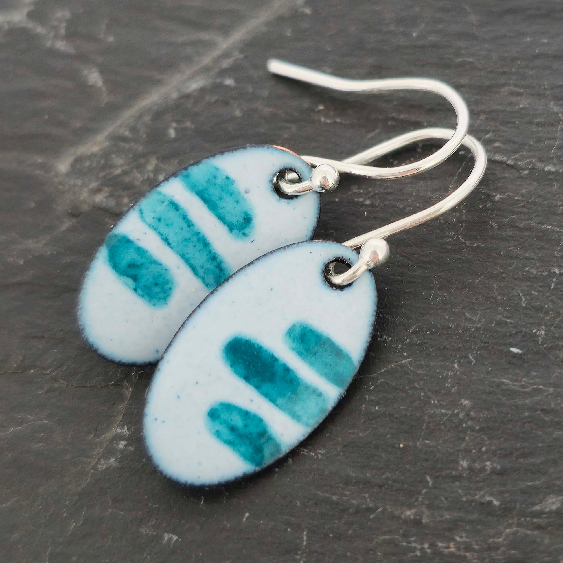 Pair of oval enamel earrings with teal stripe pattern on a light grey background with silver ear hooks on a slate background