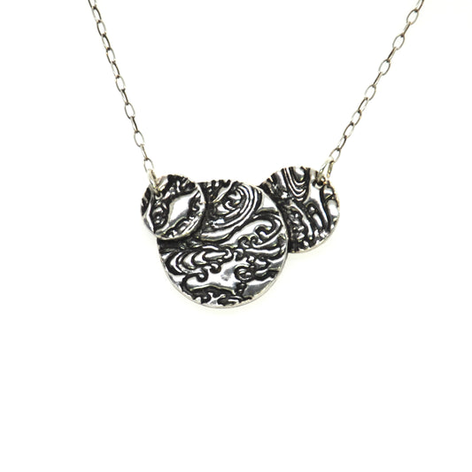 A silver necklace featuring 3 overlapping circles with a swirling water pattern on them. On a silver chain.
