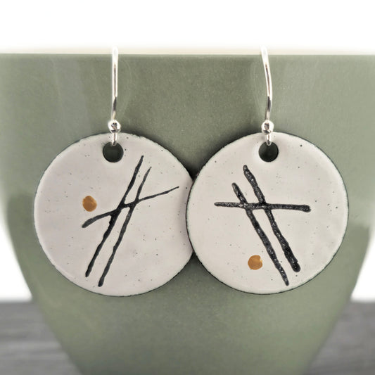 A pair of handmade white enamel round drop earrings with 3 black lines and a gold dot on each. Suspended from sterling silver ear hooks. On a pale green cup. Mijoux Jewellery.