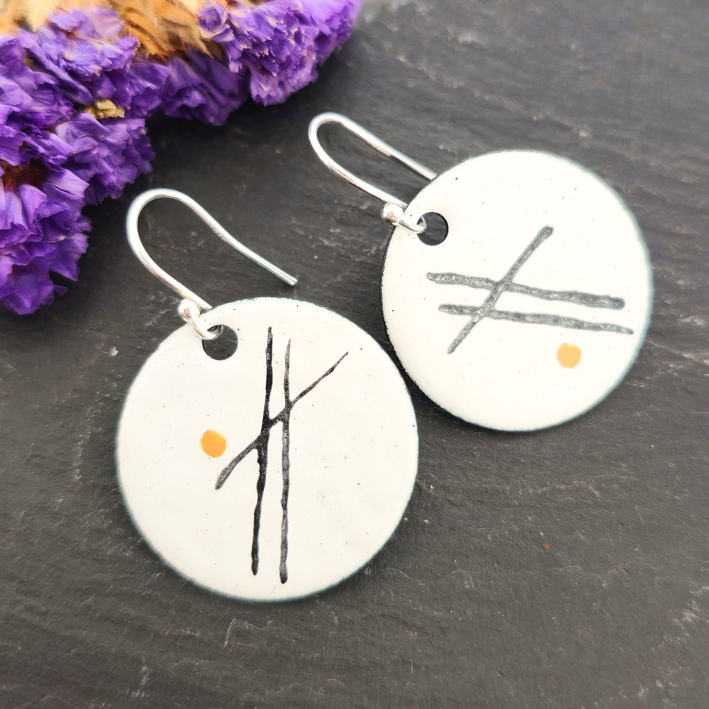A pair of handmade white enamel round drop earrings with 3 black lines and a gold dot on each. Suspended from sterling silver ear hooks. On a black background with purple flowers. Mijoux Jewellery.