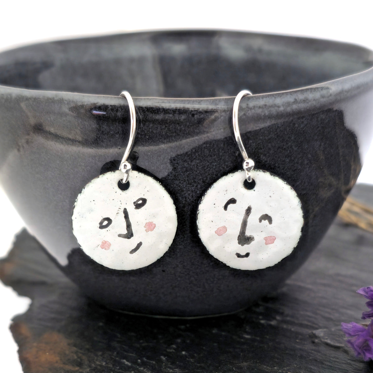 A pair of handmade enamel round drop earrings with a white background and different faces on each earrings in black and pink. On sterling silver ear hooks. Shown on a black bowl with purple flowers. Mijoux jewellery.