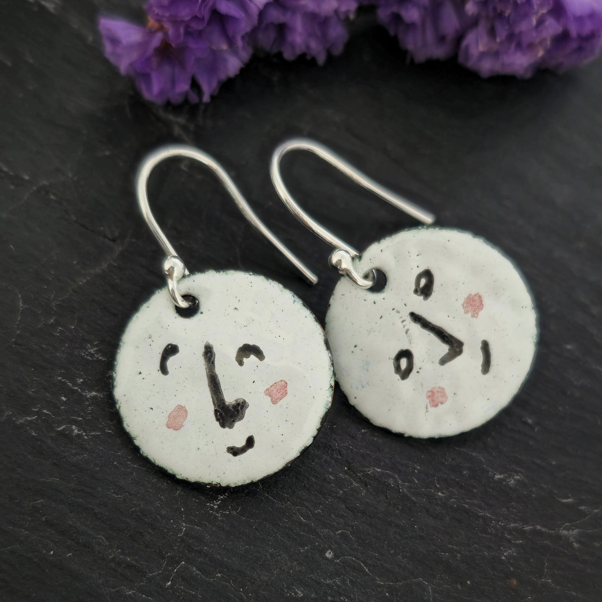 A pair of enamel round drop earrings with a white background and different faces on each earrings in black and pink. On sterling silver ear hooks. Shown on a black background with purple flowers. Mijoux jewellery.