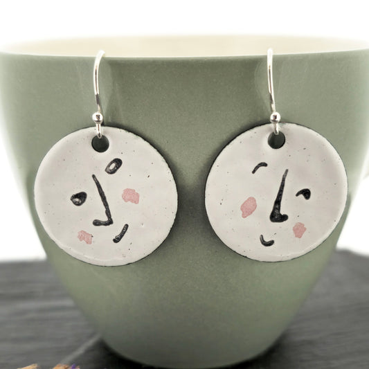 A pair of enamel round drop earrings with a white background and different faces on each earrings in black and pink. On sterling silver ear hooks. Shown on a pale green mug. Mijoux jewellery.