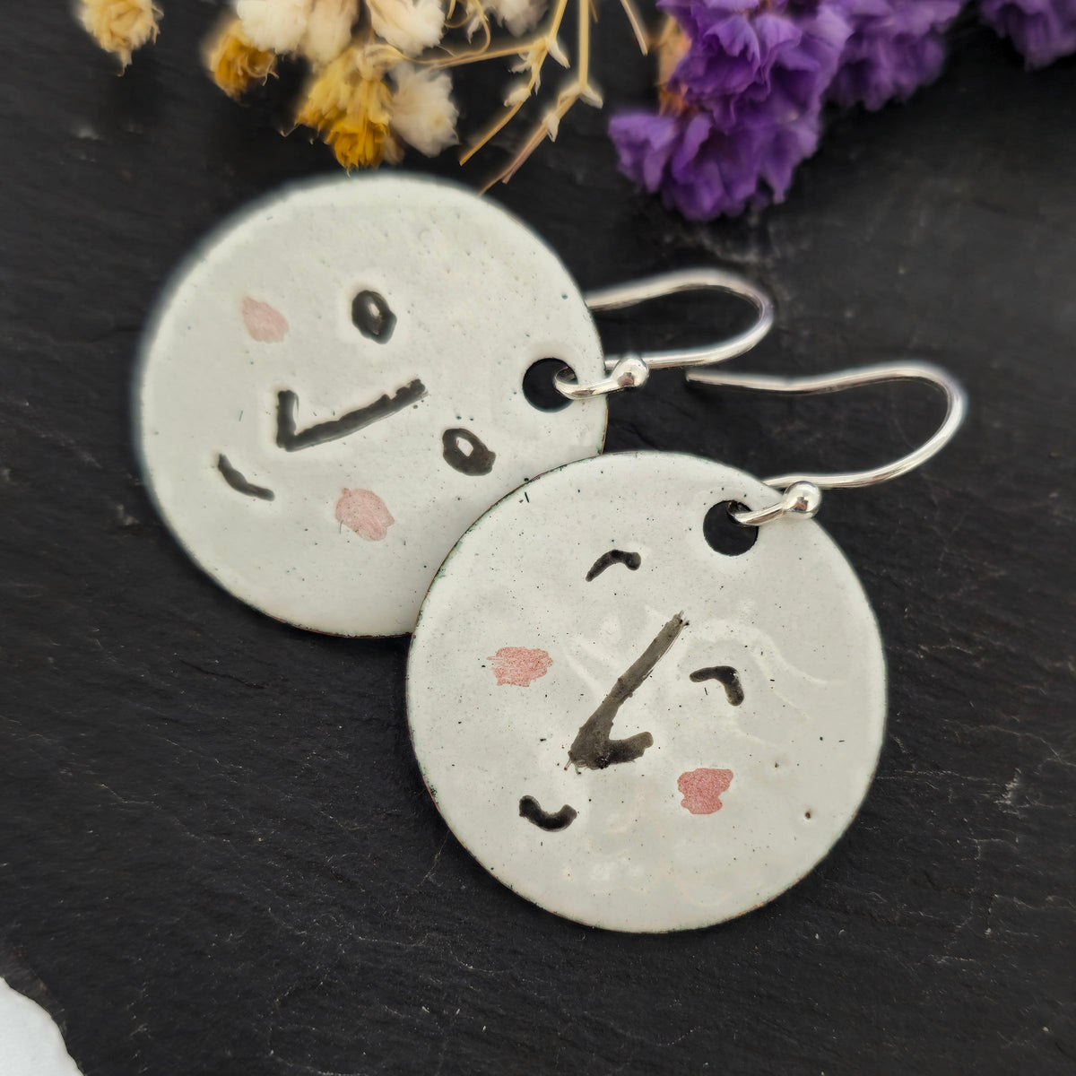 A pair of enamel round drop earrings with a white background and different faces on each earrings in black and pink. On sterling silver ear hooks. Shown on a black background with purple flowers. Mijoux jewellery.