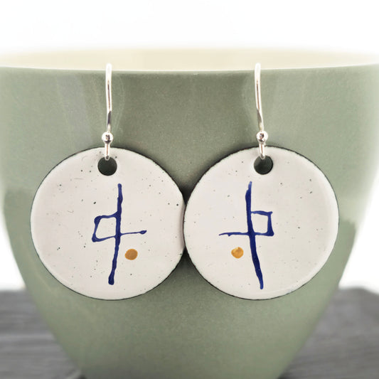 A pair of handmade white enamel round drop earrings with blue symbols and a gold dots suspended from sterling silver ear hooks. Shown on a pale green cup. Mijoux jewellery.