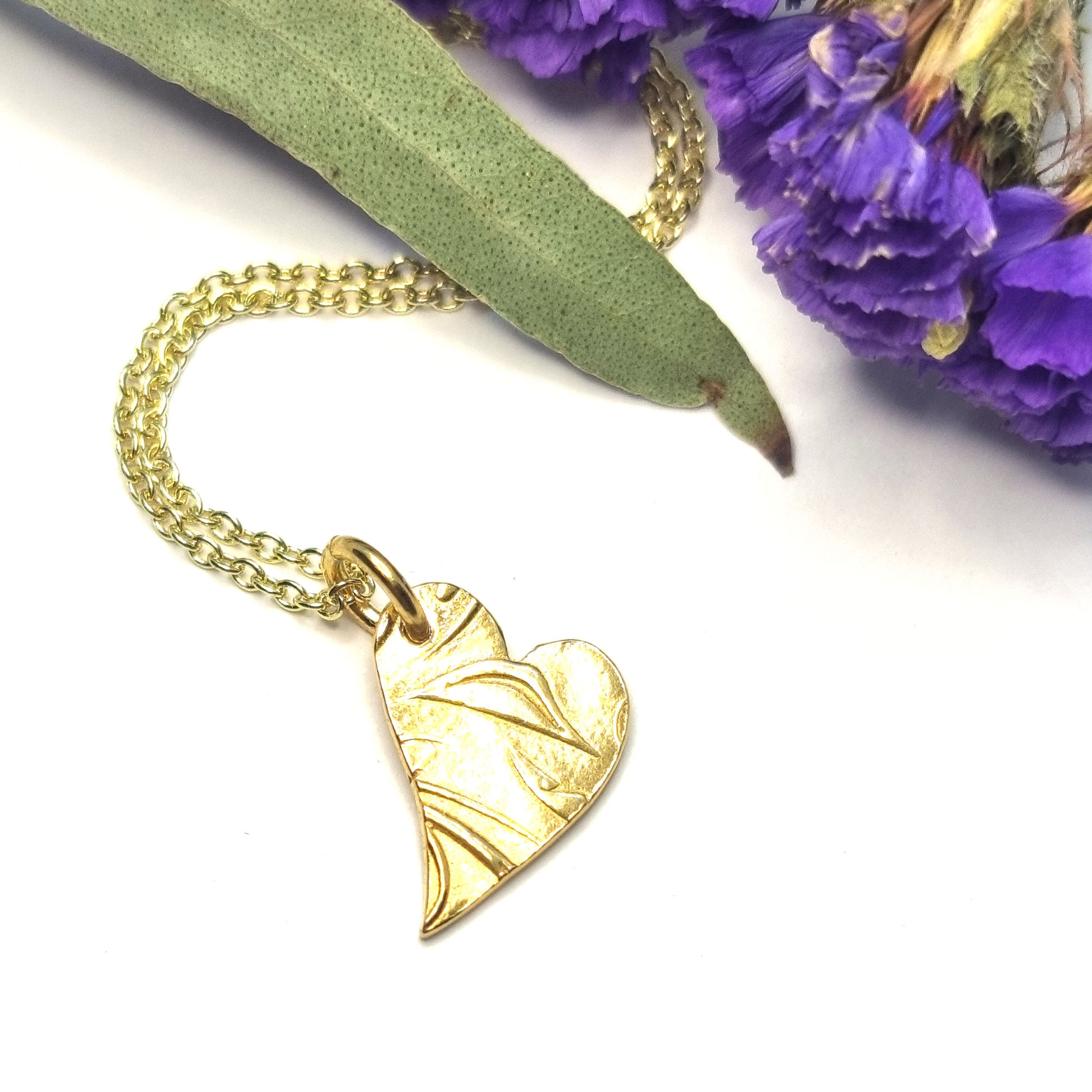 Yellow gold vermeil asymmetrical heart pendant with a leaf & vine design on a yellow gold vermeil chain. Pictured with flowers.