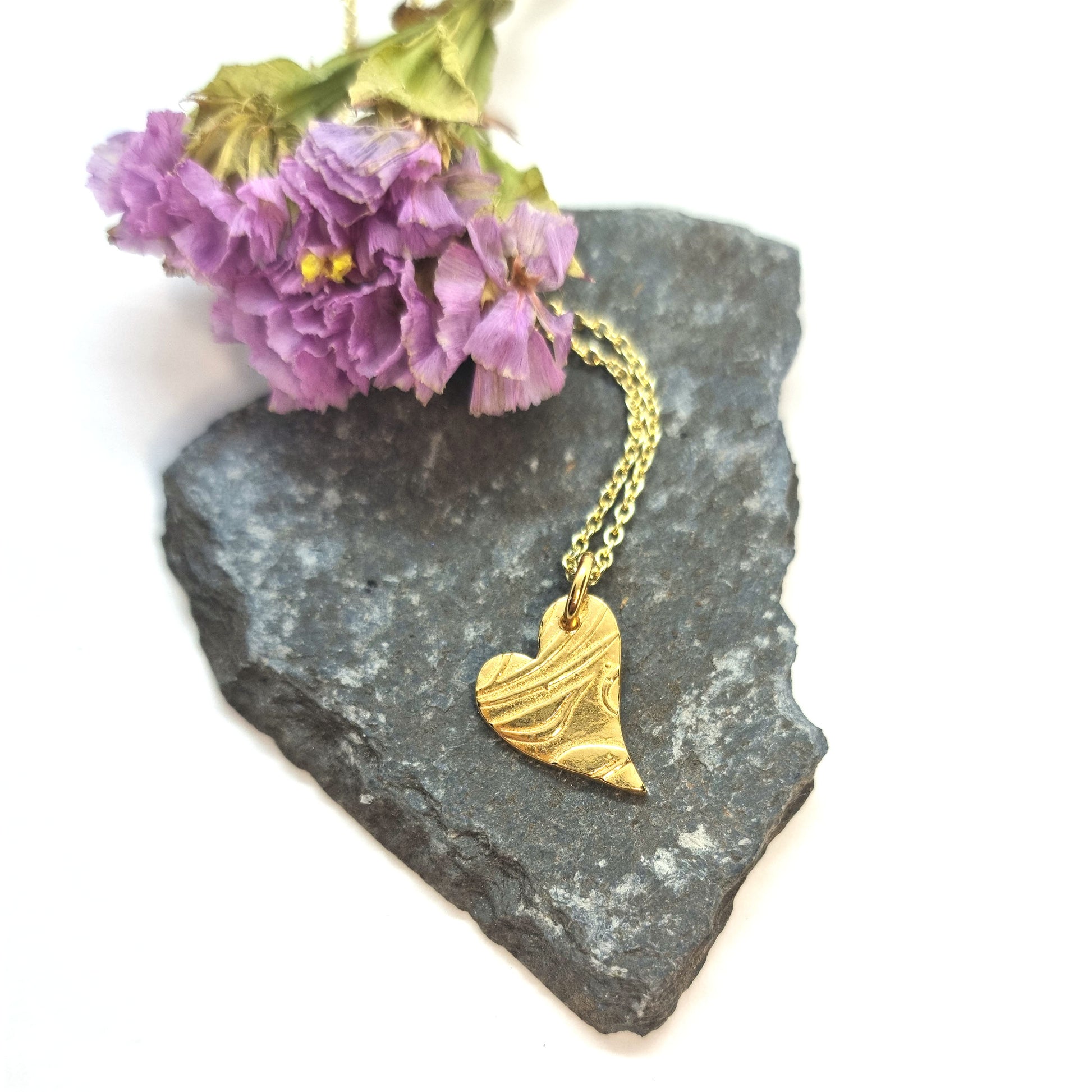 Yellow gold vermeil asymmetrical heart pendant with a leaf & vine design on a yellow gold vermeil chain. Pictured on a stone with flowers.