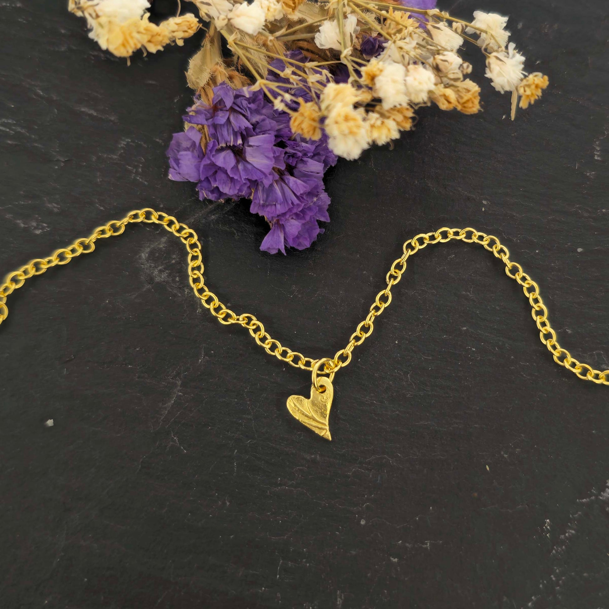 Gold bracelet with a textured asymmetric heart-shaped charm on a black background with dried flowers.