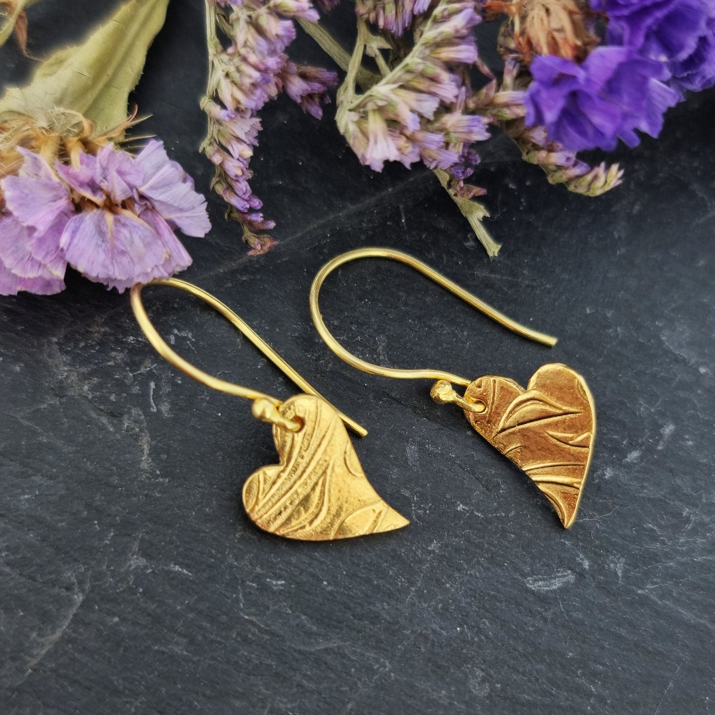 Yellow gold vermeil drop earrings featuring an asymmetrical heart with a leaf vine pattern suspended from an ear wire. Pictured with flowers.