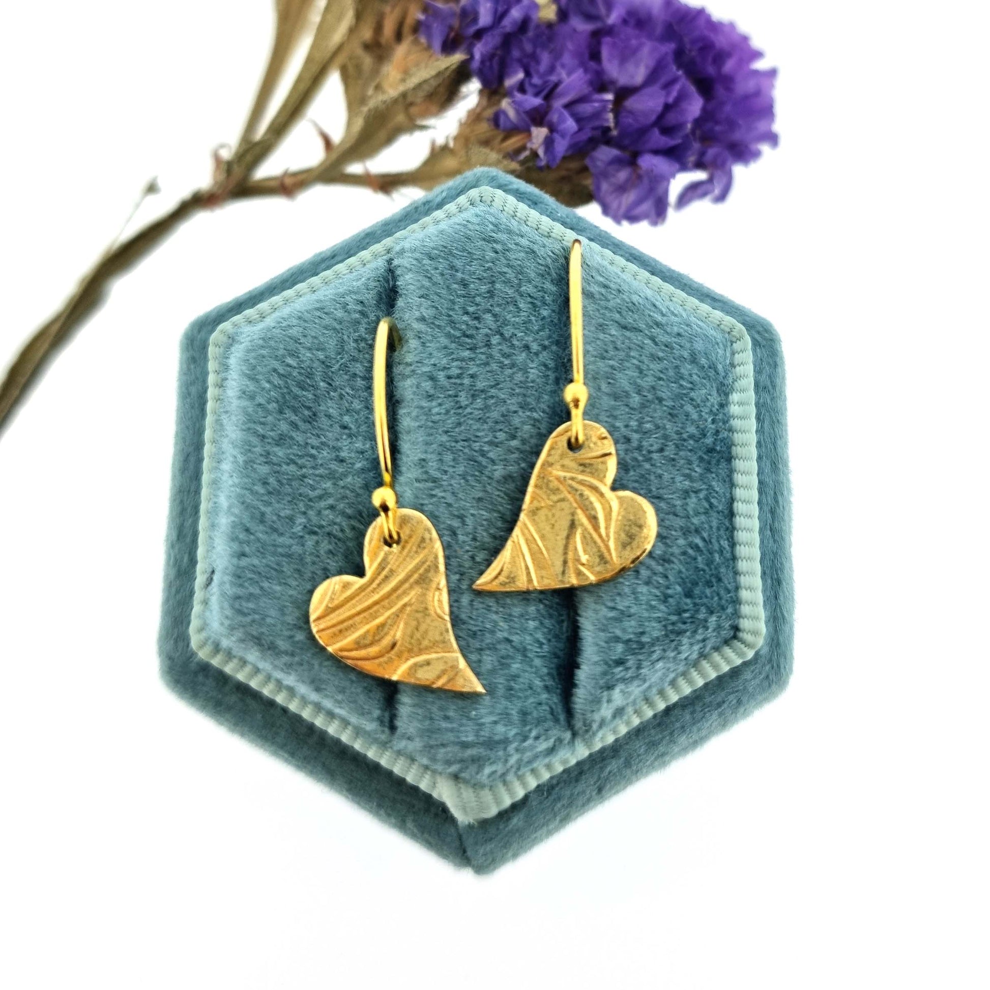 Yellow gold vermeil drop earrings featuring an asymmetrical heart with a leaf vine pattern suspended from an ear wire. Pictured in a jewellery box with flowers.