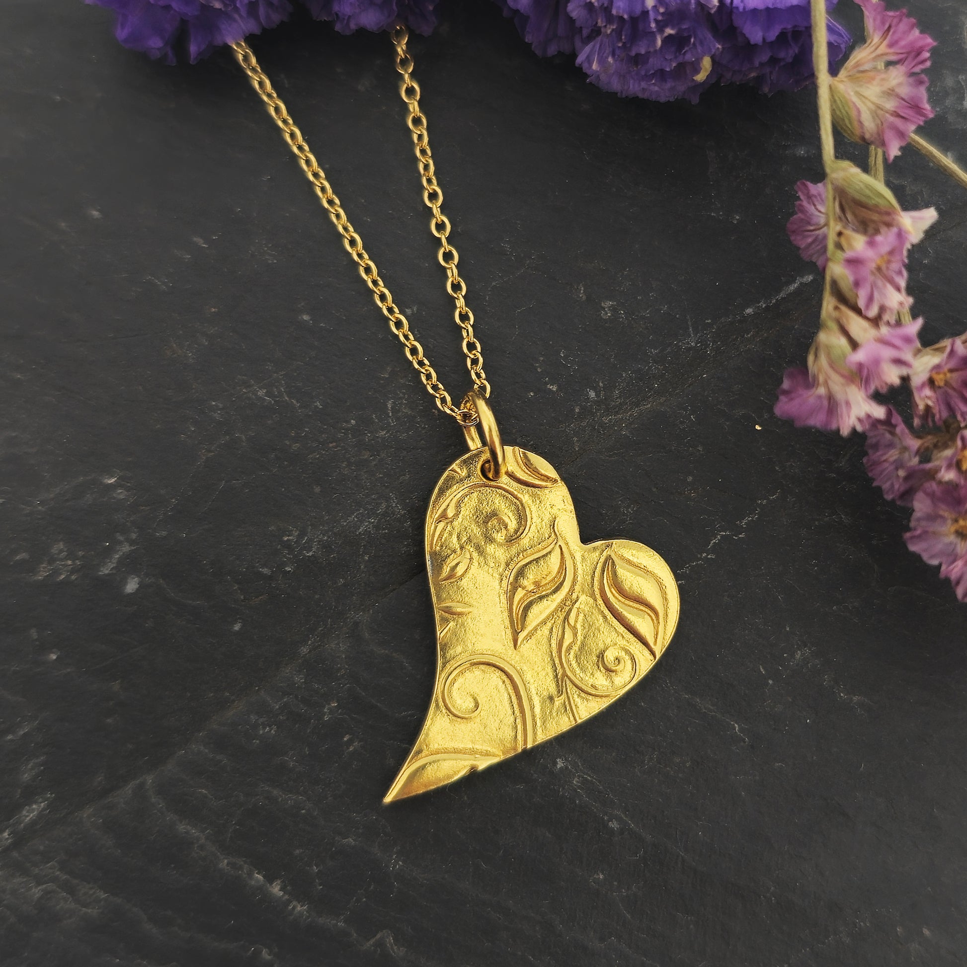 Yellow gold vermeil asymmetrical heart pendant with a leaf & vine design on a yellow gold vermeil chain. Large. Shown on slate with flowers.