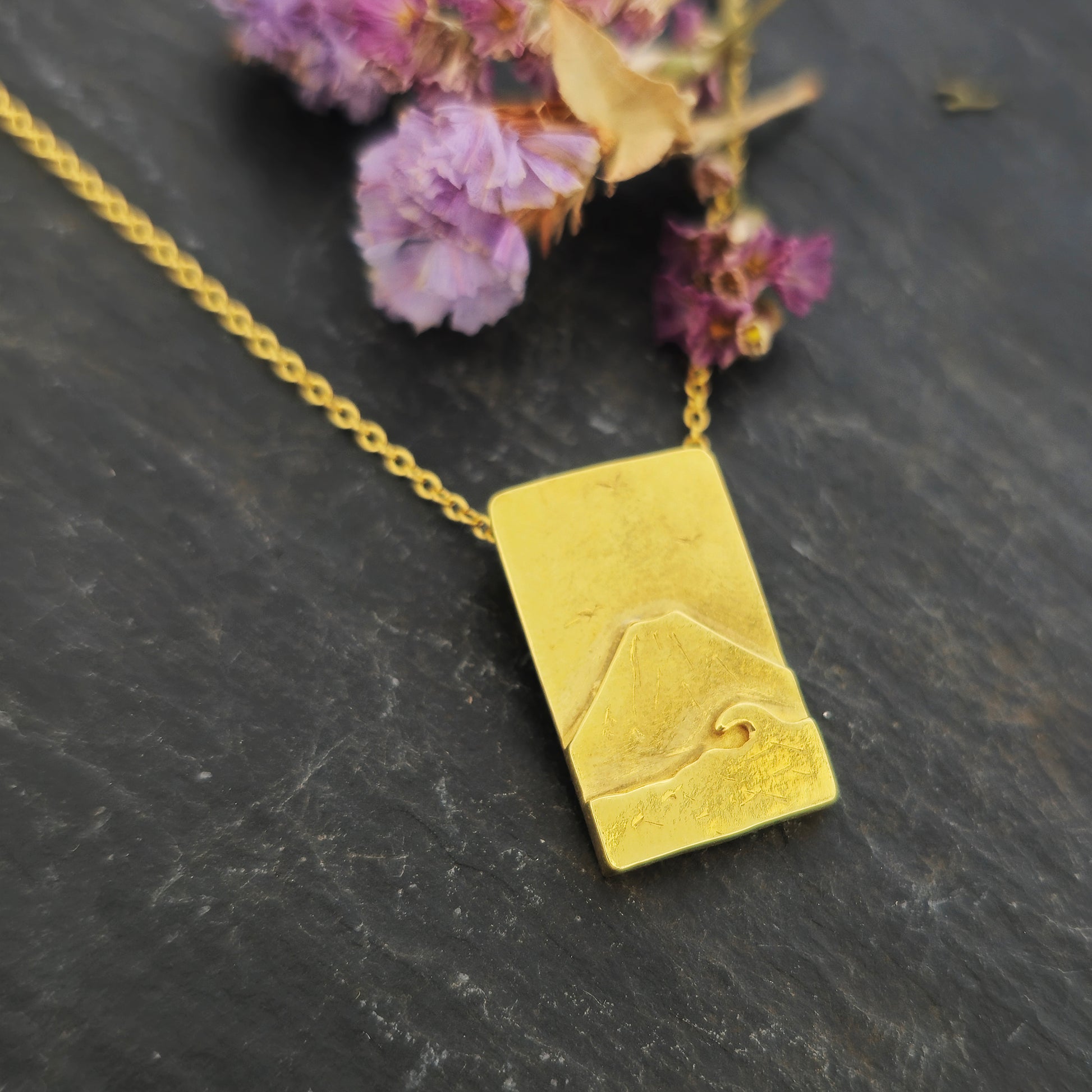 A rectangular yellow gold vermeil pendant featuring a mountain and sea scene. The pendant has an invisible bail and is on a yellow gold vermeil chain. Shown on slate with flowers.