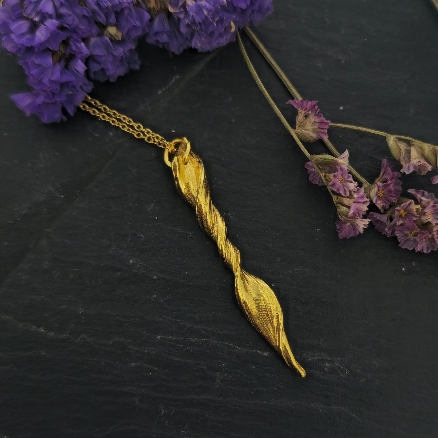 A yellow gold vermeil twist pendant suspended on a yellow gold vermeil chain. Shown on slate with flowers.