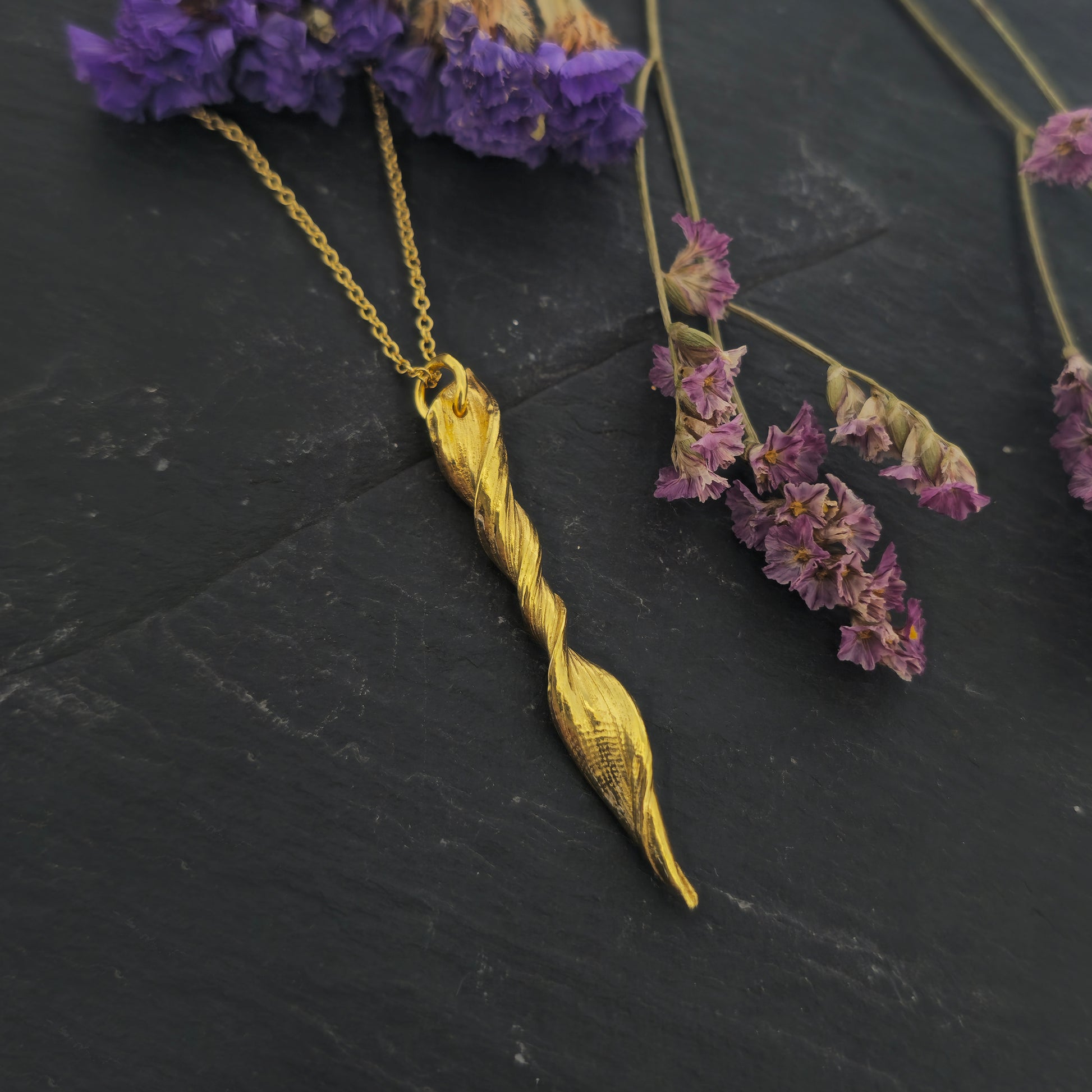 A yellow gold vermeil twist pendant suspended on a yellow gold vermeil chain. Shown on slate with flowers.